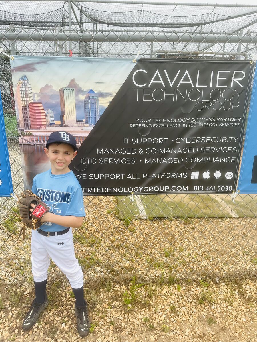 A Keystone Little League player standing in front of a Cavalier Technology Group sponsor banner on the outfield fence